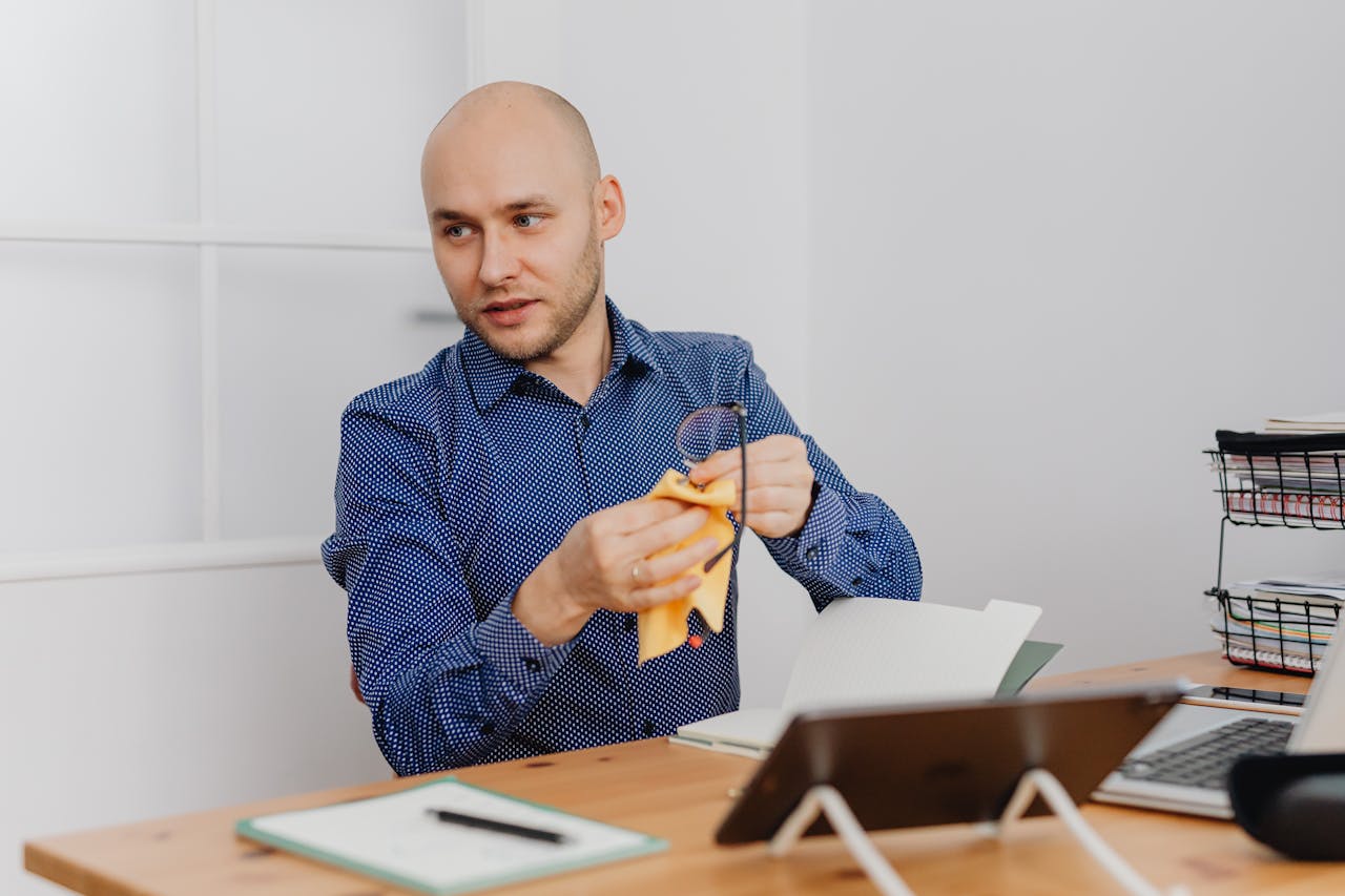 Bald man in a blue shirt cleaning eyeglasses at his office desk, emphasizing work setup and organization.